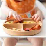 A baby in a high chair reaches for a Bumkins Silicone Grip Dish: Wood Grain with sections and a suction base, filled with small snacks.