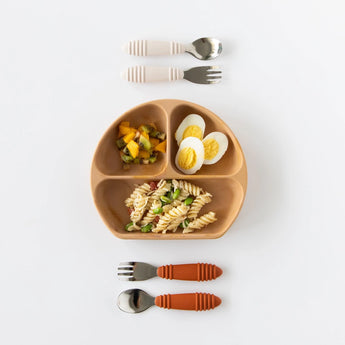 Bumkins Silicone Grip Dish in Wood Grain with pasta, eggs, and fruit salad, plus toddler utensils on a light background.