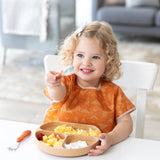 A child with curly hair and an orange bib holds a spoon at the table with a Bumkins Silicone Grip Dish: Wood Grain full of food.