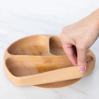 Hand lifting a Bumkins Silicone Grip Dish: Wood Grain from a marble surface, crafted with food-safe silicone.