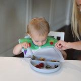 A toddler self-feeds fruit from a Bumkins Silicone Grip Dish (Gray) in a high chair, wearing a themed bib, with an adult assisting.