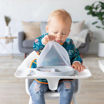 A toddler in a colorful bib eagerly opens a Bumkins Silicone Grip Dish with Lid (3 Section): Gray, designed for toddlers. The child is focused on the dishs suction base, while blurred living room furniture appears in the background.