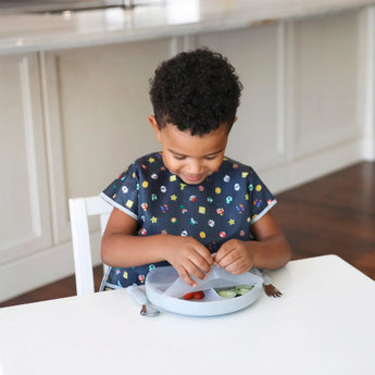 A toddler wearing a bib self-feeds from a Bumkins Silicone Grip Dish with Lid (3 Section), Gray, featuring a secure suction base.