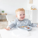 A child happily self-feeds with a Bumkins Silicone Grip Dish; gray, securely on the table in a bright room.