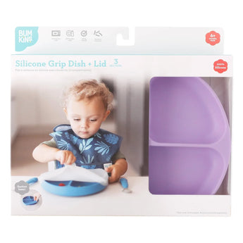 A toddler with a bib uses a blue silicone dish; packaging shows Bumkins Lavender Silicone Grip Dish with Lid, made of food-safe silicone.