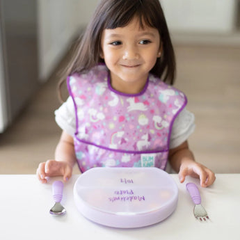 A child with a purple bib smiles at a table set with Bumkins Silicone Grip Dish in Lavender, complete with matching utensils.