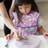 A child uses a Lavender Silicone Grip Dish with Lid by Bumkins to write, assisted by an adult.