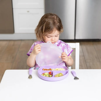 A child in a purple bib opens a Bumkins Lavender Silicone Grip Dish with Lid at a table, near a fork and spoon.