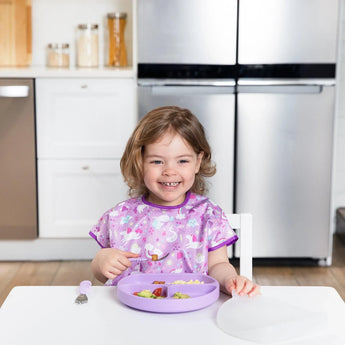 A smiling toddler in a purple bib sits at the kitchen table with a Bumkins Silicone Grip Dish in Lavender near a stainless steel fridge.