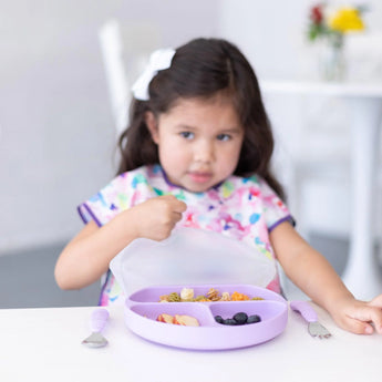 A young girl holds the Silicone Grip Dish with Lid (3 Section) in Lavender by Bumkins, filled with snacks and utensils, at a table.