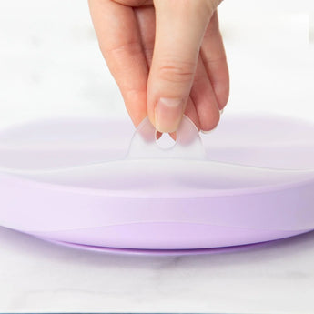 A hand lifts the translucent lid from a Bumkins Lavender Silicone Grip Dish with 3 sections.