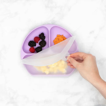 A hand lifts the lid of Bumkins Lavender Silicone Grip Dish with compartments holding berries, orange puree, and cooked pasta.