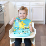 A toddler smiles in a high chair, wearing a bib, holding a spoon beside her Bumkins Silicone Grip Dish with Lid (5 Section): Blue.