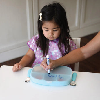 A child writes on the blue Bumkins Silicone Grip Dish with Lid, guided by an adults hand; its a food-safe silicone canvas.