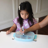 A young girl uses the CPSIA compliant *Bumkins Silicone Stretch Lid for Grip Dish*, an adult helps her write while a fork and spoon rest nearby.