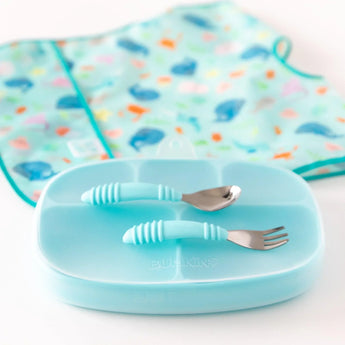 Bumkins Blue Silicone Grip Dish with Lid shown with matching fork and spoon in front of a colorful baby bib on a white surface.