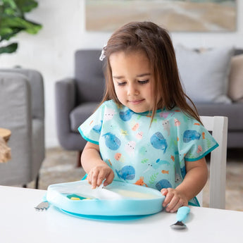 A young child enjoys a meal using Bumkins Blue Silicone Grip Dish with 5 sections, in a cozy living room setting.