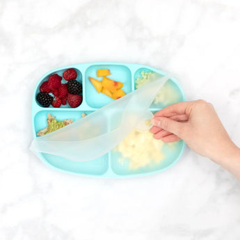 A hand lifts Bumkins CPSIA-compliant Silicone Stretch Lid off a blue Grip Tray, revealing its five sections filled with berries, diced peaches, rice, and vegetables. The tray sits elegantly on a white marble surface.