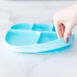 A hand holds a blue Bumkins Silicone Grip Dish with Lid (5 Section) made of food-safe silicone, resting on a light marble surface.