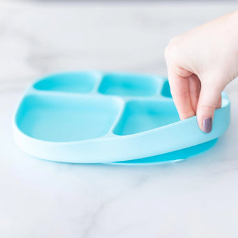 A hand holds a blue Bumkins Silicone Grip Dish with Lid (5 Section) made of food-safe silicone, resting on a light marble surface.