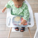 In a high chair, a toddler in a green animal-print bib reaches for a Bumkins Grip Dish with Silicone Stretch Lid beside the utensils.