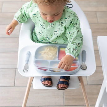 In a high chair, a toddler in a green animal-print bib reaches for a Bumkins Grip Dish with Silicone Stretch Lid beside the utensils.