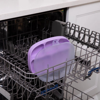 A Lavender Silicone Grip Dish with Lid by Bumkins, made for toddler portions, rests in a dishwashers top rack. Food-safe and versatile.