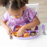 A curly-haired child enjoys toddler portions from a lavender Bumkins Silicone Grip Dish, wearing a matching bib at a white table.