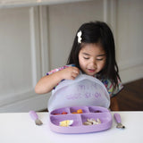 A child with long dark hair sits at a table, excitedly opening the lid on a Bumkins Silicone Grip Dish with Lid (5 Section) in Lavender, designed for toddler portions. A fork and spoon are neatly placed beside the dish. The child wears a colorful shirt and has a charming hair accessory.