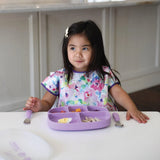 A young girl sits at a table with a Bumkins Lavender Silicone Grip Dish, wearing a floral bib, looking to the side.