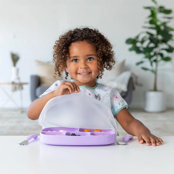 A child smiles, opening a purple Bumkins lunchbox with fruit inside, sitting at a table with a colorful Grip Tray in a bright room.