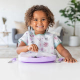 A smiling child with a bib plays with a purple Bumkins silicone stretch lid for Grip Dish in a bright room, blurred background.