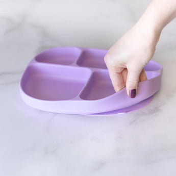 A hand lifts a Bumkins Silicone Grip Dish with Lid (5 Section), showcasing its food-safe, flexible Lavender design.