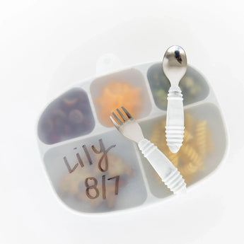 A white fork and spoon rest on a Bumkins silicone stretch lid for Grip Dish, featuring Lily and 817, covering an assorted lunch.