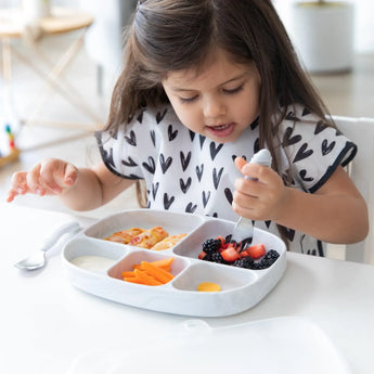 A young girl enjoys snacks from a Bumkins Marble Silicone Grip Dish with Lid at a white table, filled with berries, cheese, and sauce.