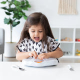 A young girl joyfully eats from a Bumkins Marble Silicone Grip Dish with Lid at the table in a heart-patterned shirt.