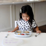 A young girl with a hair clip smiles at a meal on her Bumkins Silicone Grip Dish from the Marble collection in a heart-patterned shirt.