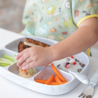 A child wearing a patterned bib reaches for food on a Bumkins Silicone Grip Dish with Lid: Marble, secure with its suction base.