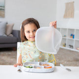 Child smiling while lifting the lid of a Bumkins Silicone Grip Dish with Lid (5 Section): Marble, sitting at a table with utensils.