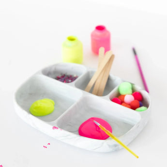 The Bumkins Marble Silicone Grip Dish with Lid (5 Section) keeps neon paints, pom-poms, glitter, and sticks tidy on the white background.