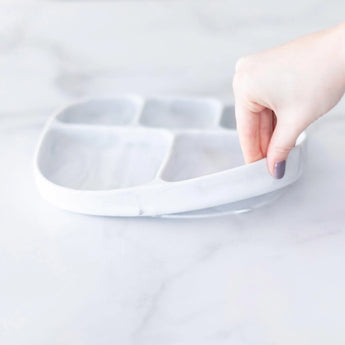 A hand lifts a Bumkins Marble Silicone Grip Dish (5 sections) with a suction base on a white surface.