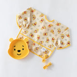 Disney Little Ones Gift Bundle by Bumkins: Yellow bib and Winnie the Pooh bowl, fork, and spoon set on a light background.