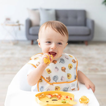 A baby in a high chair uses a spoon with a Silicone Grip Dish, wearing an animal-print bib. A cozy sofa and Winnie The Pooh Gift Bundle by Bumkins are nearby.