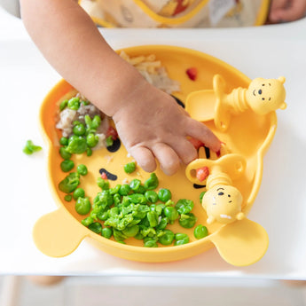 A child reaches for peas from a Bumkins yellow Winnie The Pooh dish, making mealtime fun and mess-free.