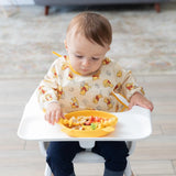 A baby in a high chair, wearing a Bumkins patterned bib, enjoys snacks from a yellow tray with their Disney Little Ones Gift Bundle.