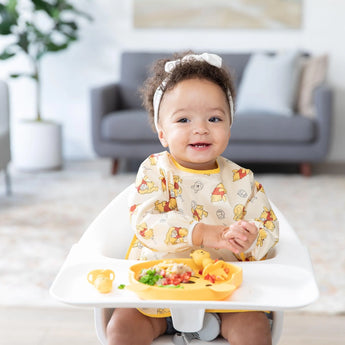 A baby in a high chair wears Disney Little Ones bib & headband, enjoying a meal on Bumkins Silicone Grip Dish; sofa & plant behind.