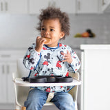 In a bright kitchen, a toddler wearing a Disney Little Ones shirt enjoys snacks from a Bumkins Silicone Grip Dish while in the high chair.