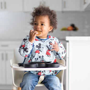 In a bright kitchen, a toddler wearing a Disney Little Ones shirt enjoys snacks from a Bumkins Silicone Grip Dish while in the high chair.