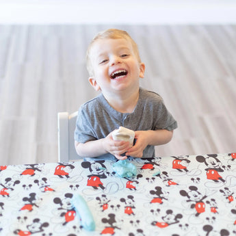 A toddler on a Mickey Mouse tablecloth holds play dough, with a Bumkins Disney Little Ones Silicone Grip Dish nearby.