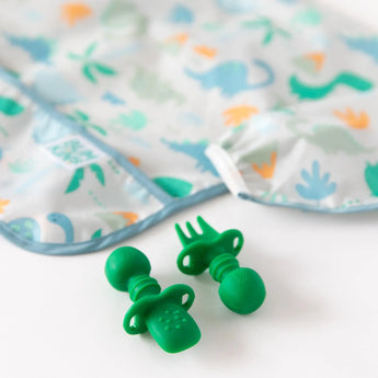 On a white table, Bumkins Ultimate Gift Set - Dinosaurs & Blue Tropic adds playful charm with a bib, dish, and Chewtensils.